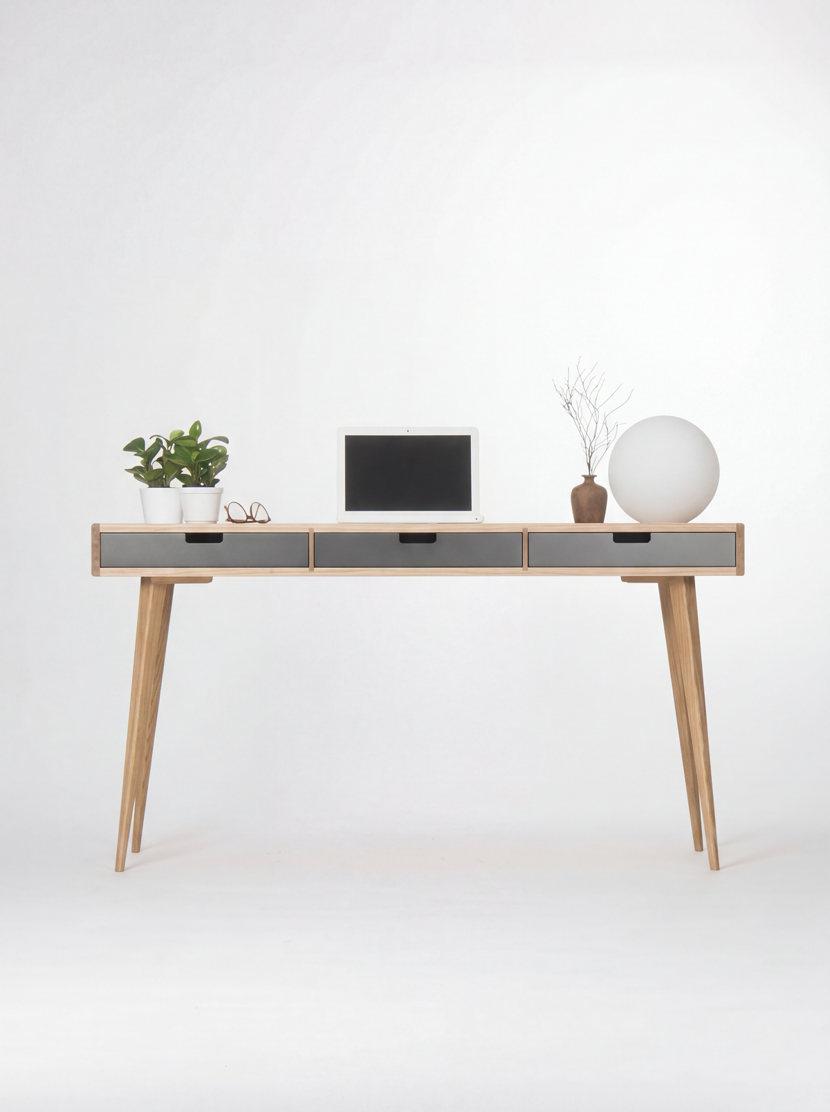 Head-on view of the HEIM 140 desk in oak wood with three black drawer fronts, a laptop, two plants and a globe lamp