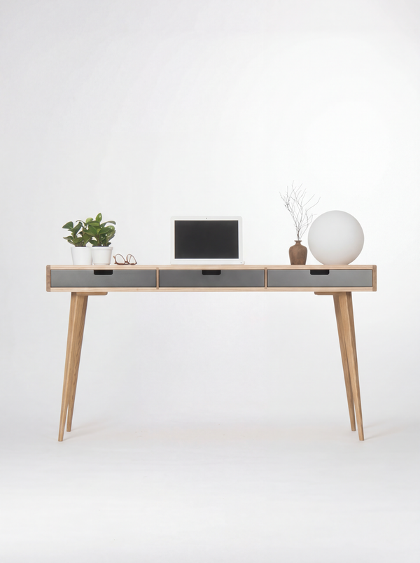 Head-on view of the HEIM 140 desk in oak wood with three black drawer fronts, a laptop, two plants and a globe lamp