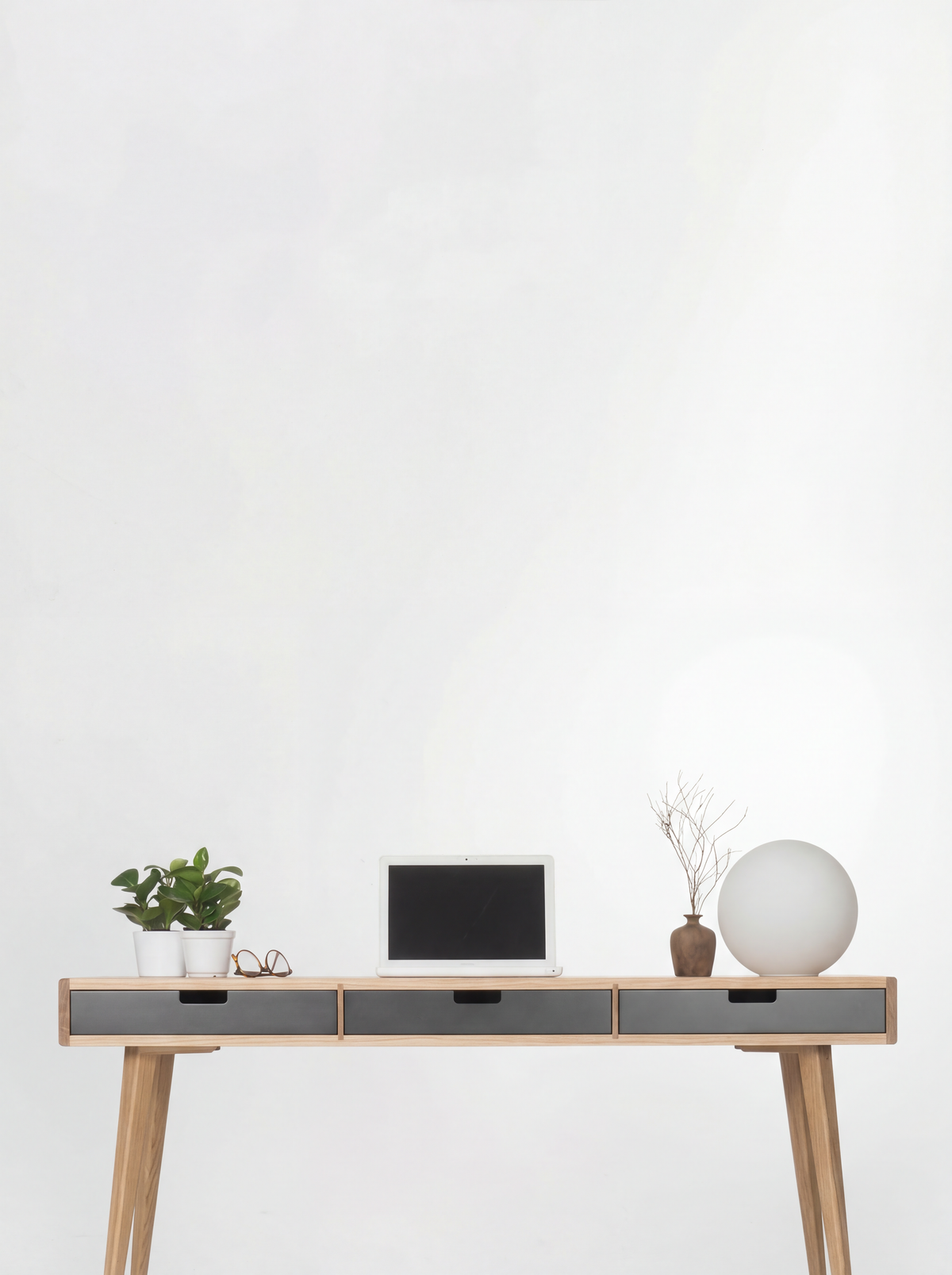 HEIM 140 desk in oak wood against a white wall, with black drawer fronts, a laptop, plants and a globe lamp