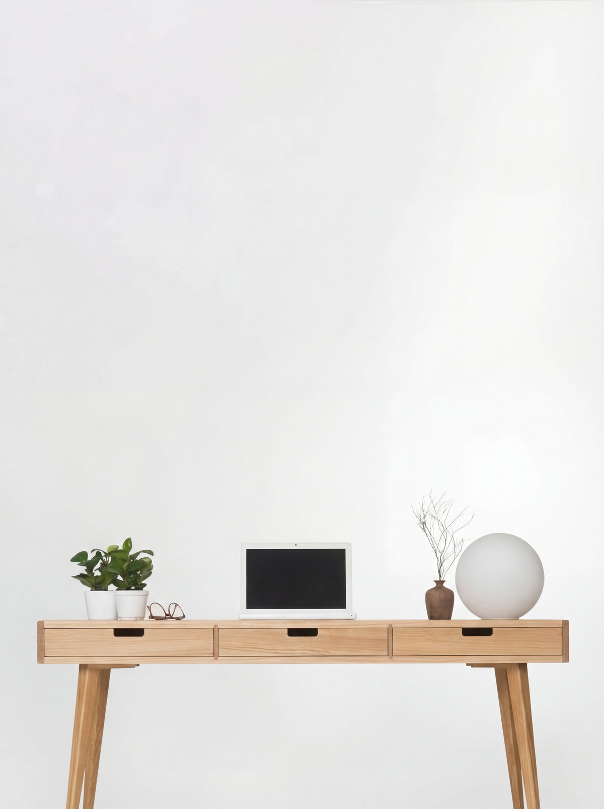 Front view of the HEIM 140 desk in oak wood, with three oak drawer fronts, a laptop, plants and a globe lamp