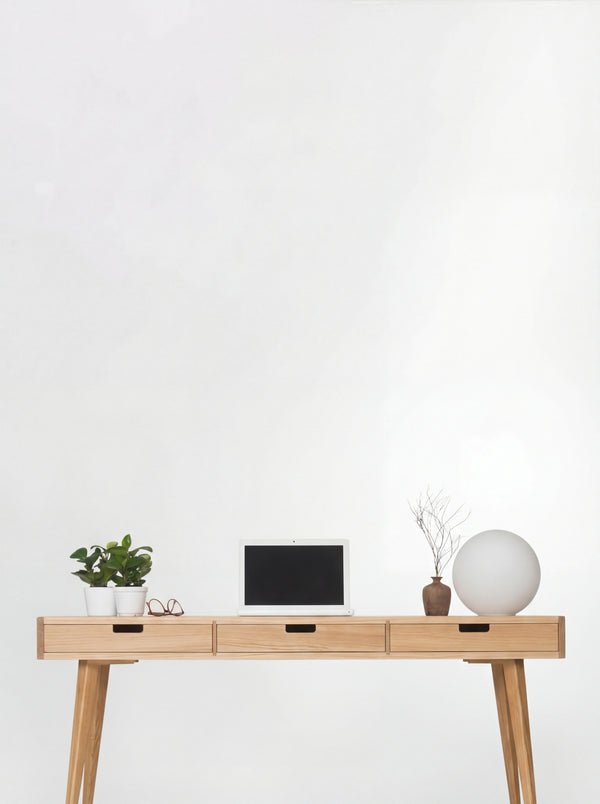 Front view of the HEIM 140 desk in oak wood, with three oak drawer fronts, a laptop, plants and a globe lamp