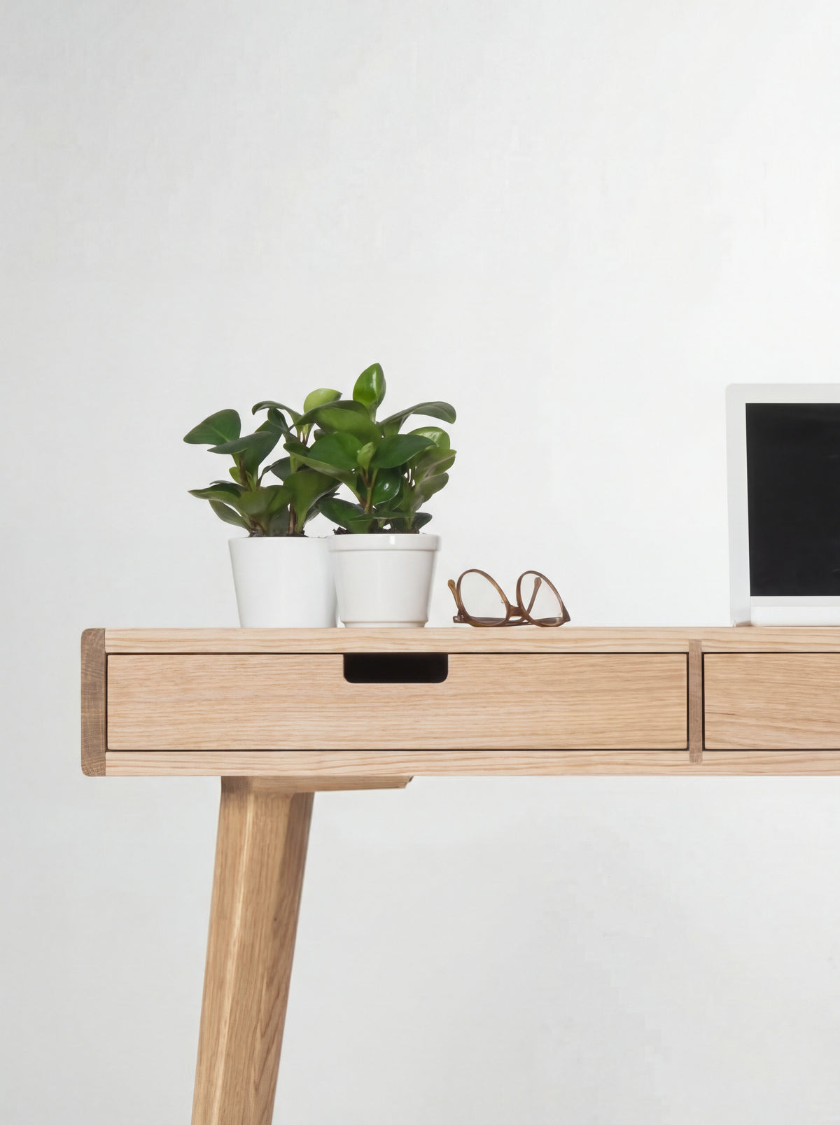 Close-up of the HEIM 140 desk in oak wood, showing the left drawer's cut-out grip with plants and glasses above