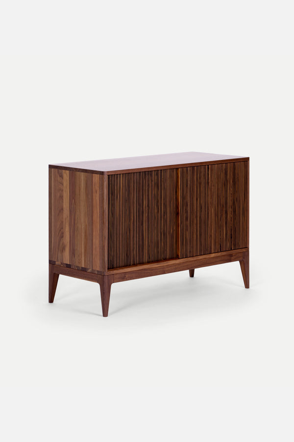 Wooden chest of drawers with a ribbed front design and tapered legs against a white background.