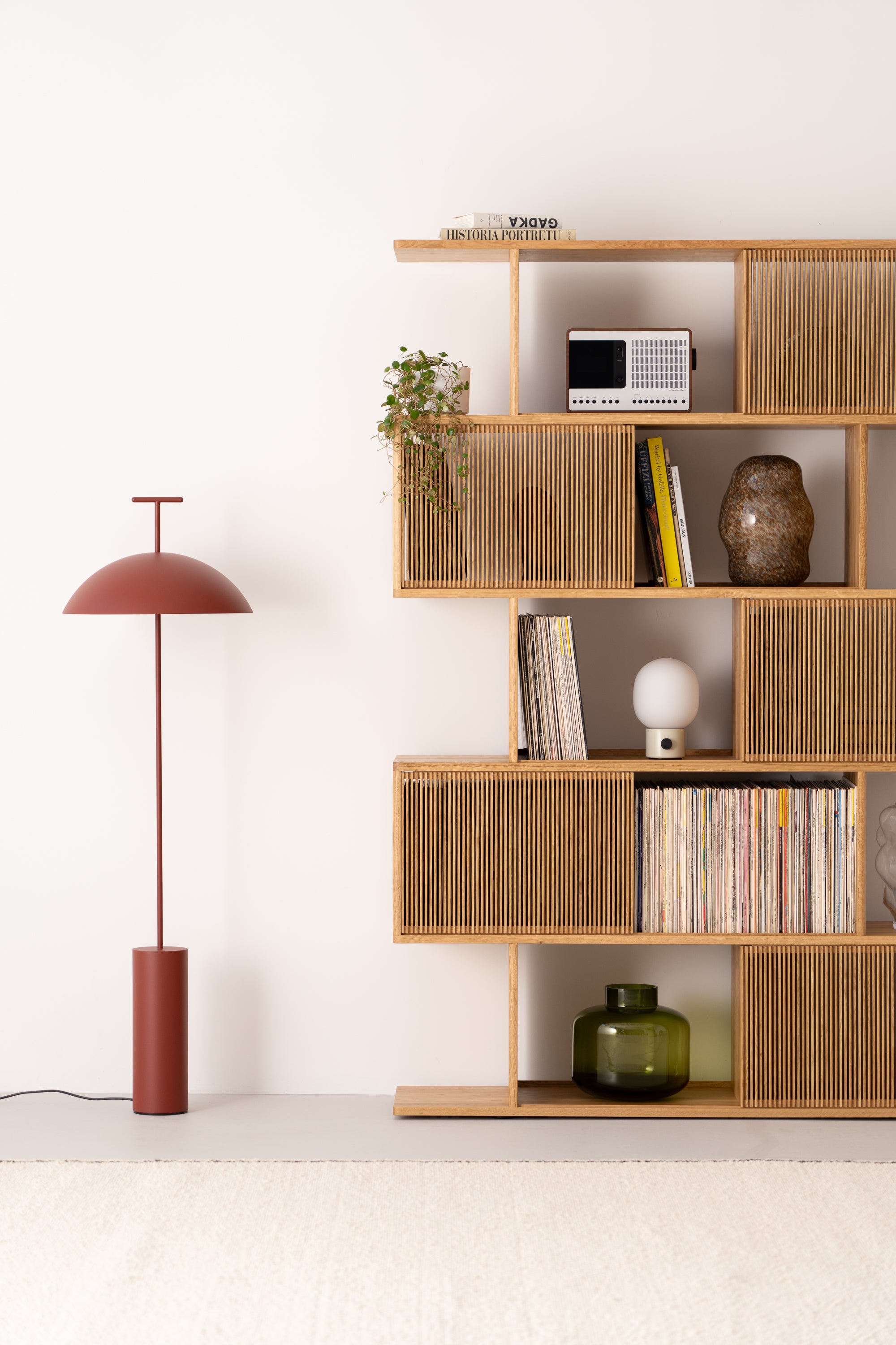 Minimal room with a wooden shelf, books, and a red floor lamp against a white wall.