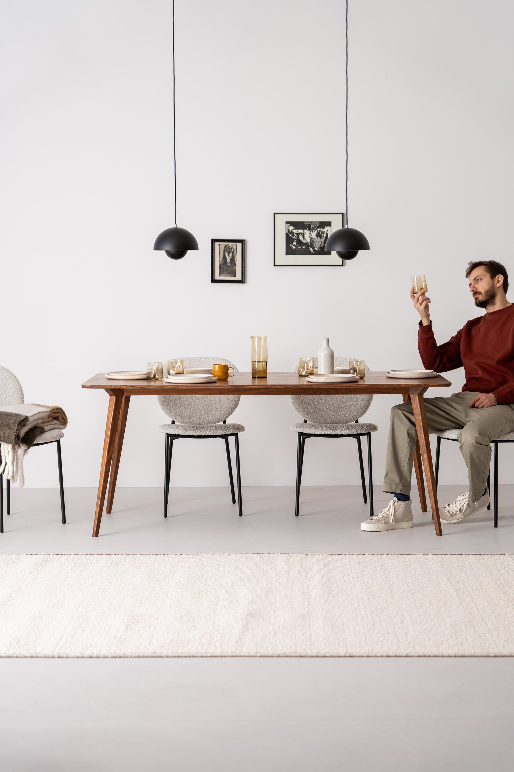 Man in a red sweater, sitting at a minimalist dining table holding a glass, with two black pendant lamps overhead.