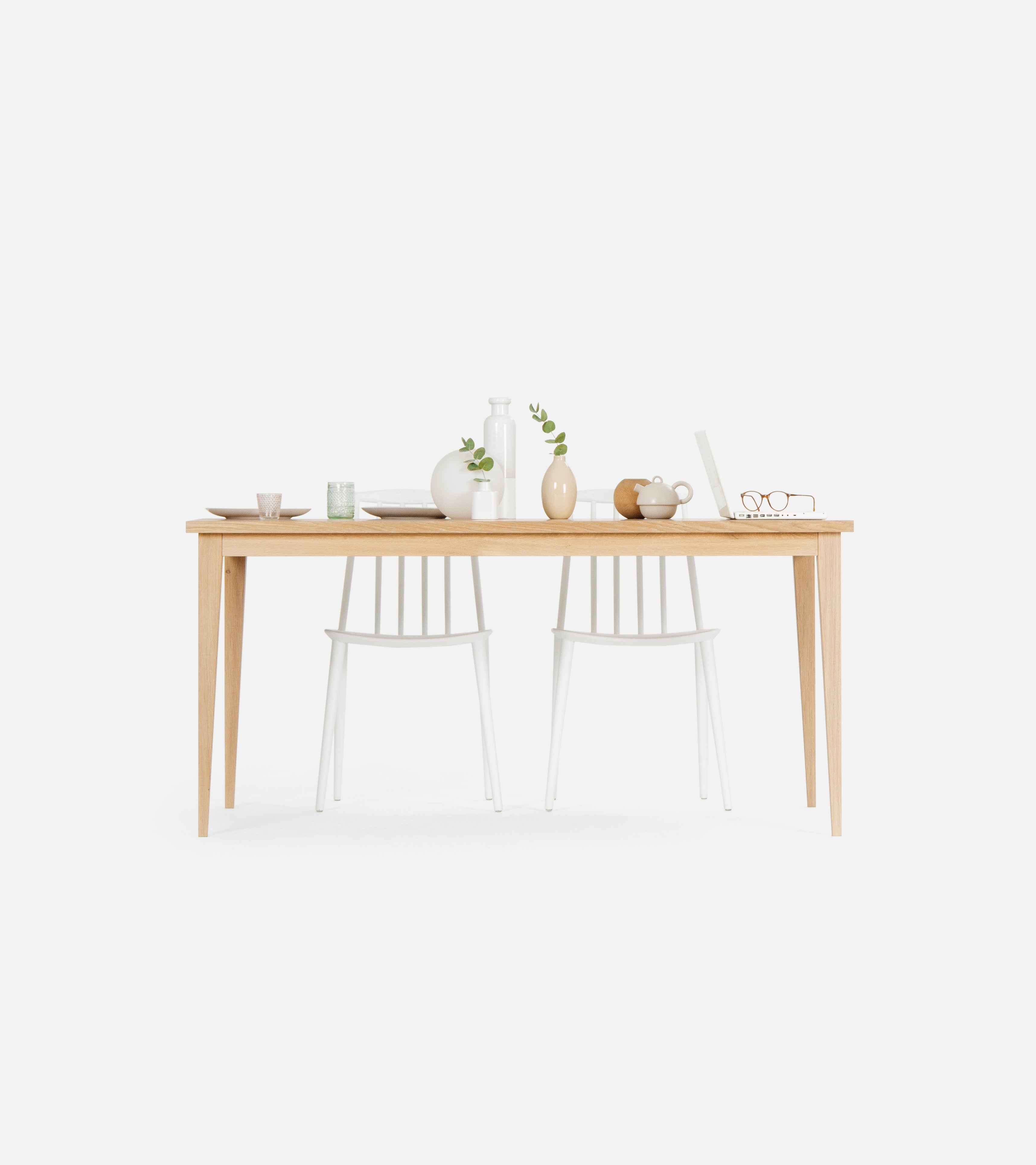 Minimalist wooden dining table with white chairs, decorated with ceramics, vases, and a pair of glasses on a book.