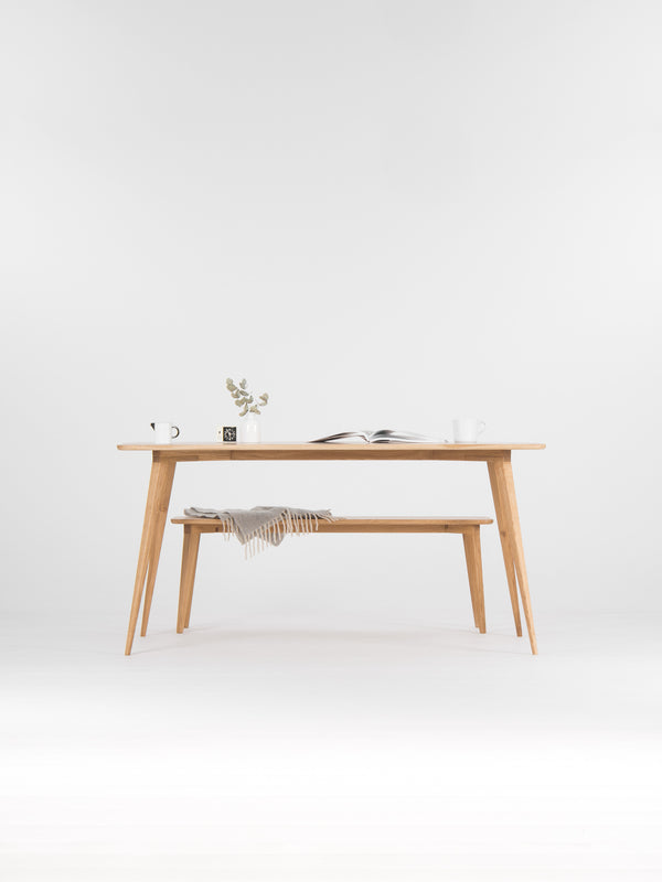 Minimalist wooden table with a plant, open book, and mugs on top; a gray blanket on the bench below.