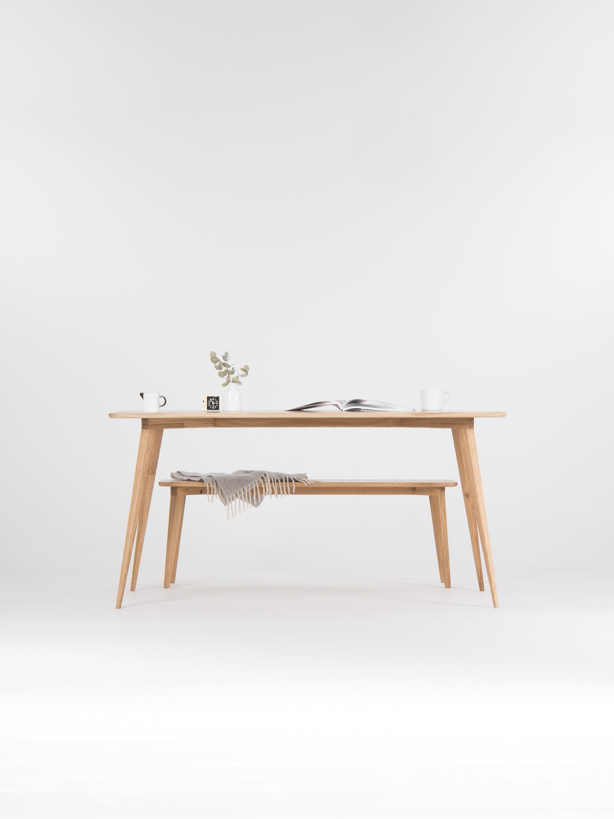 Minimalist wooden table with a plant, open book, and mugs on top; a gray blanket on the bench below