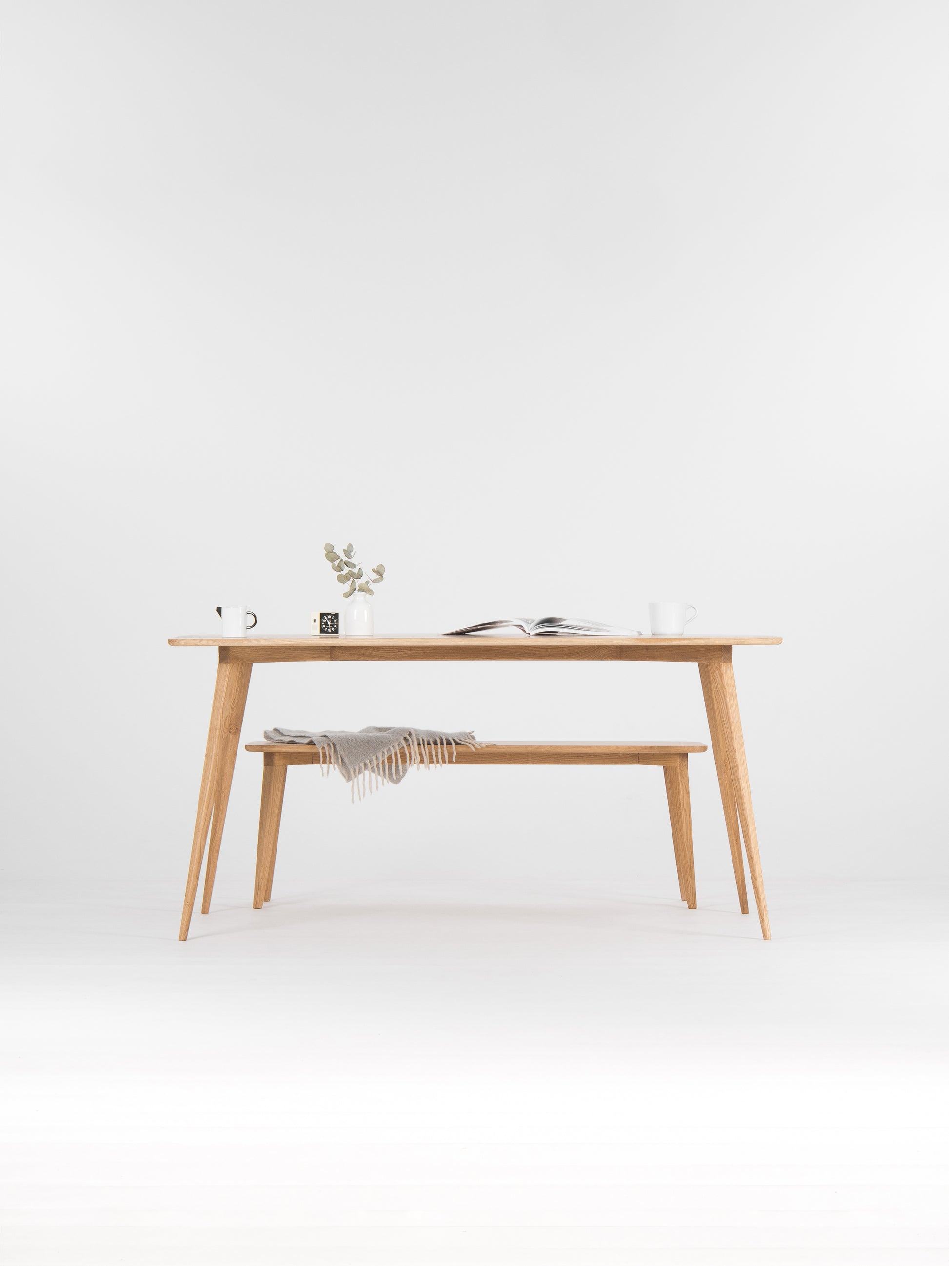 Minimalist wooden table with a plant, open book, and mugs on top; a gray blanket on the bench below.