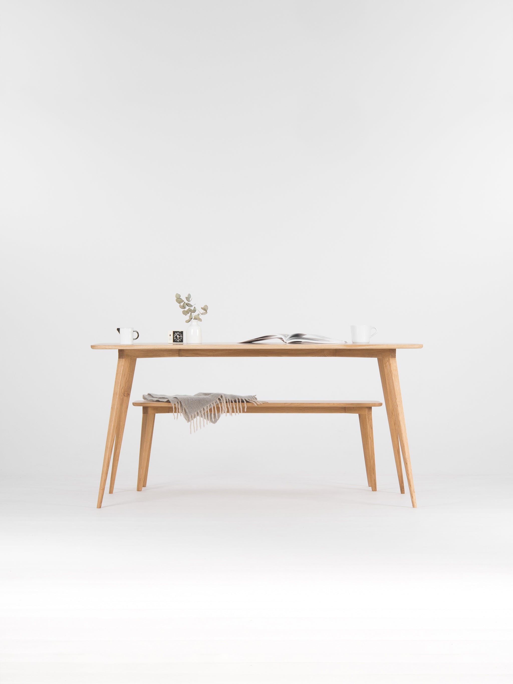 Minimalist wooden table with a plant, open book, and mugs on top; a gray blanket on the bench below.