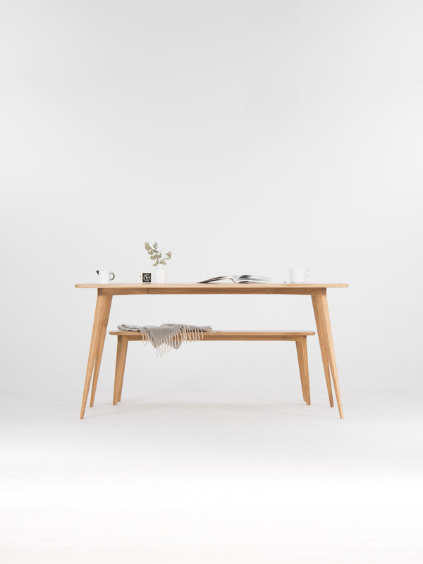 Minimalist wooden table with a plant, open book, and mugs on top; a gray blanket on the bench below.