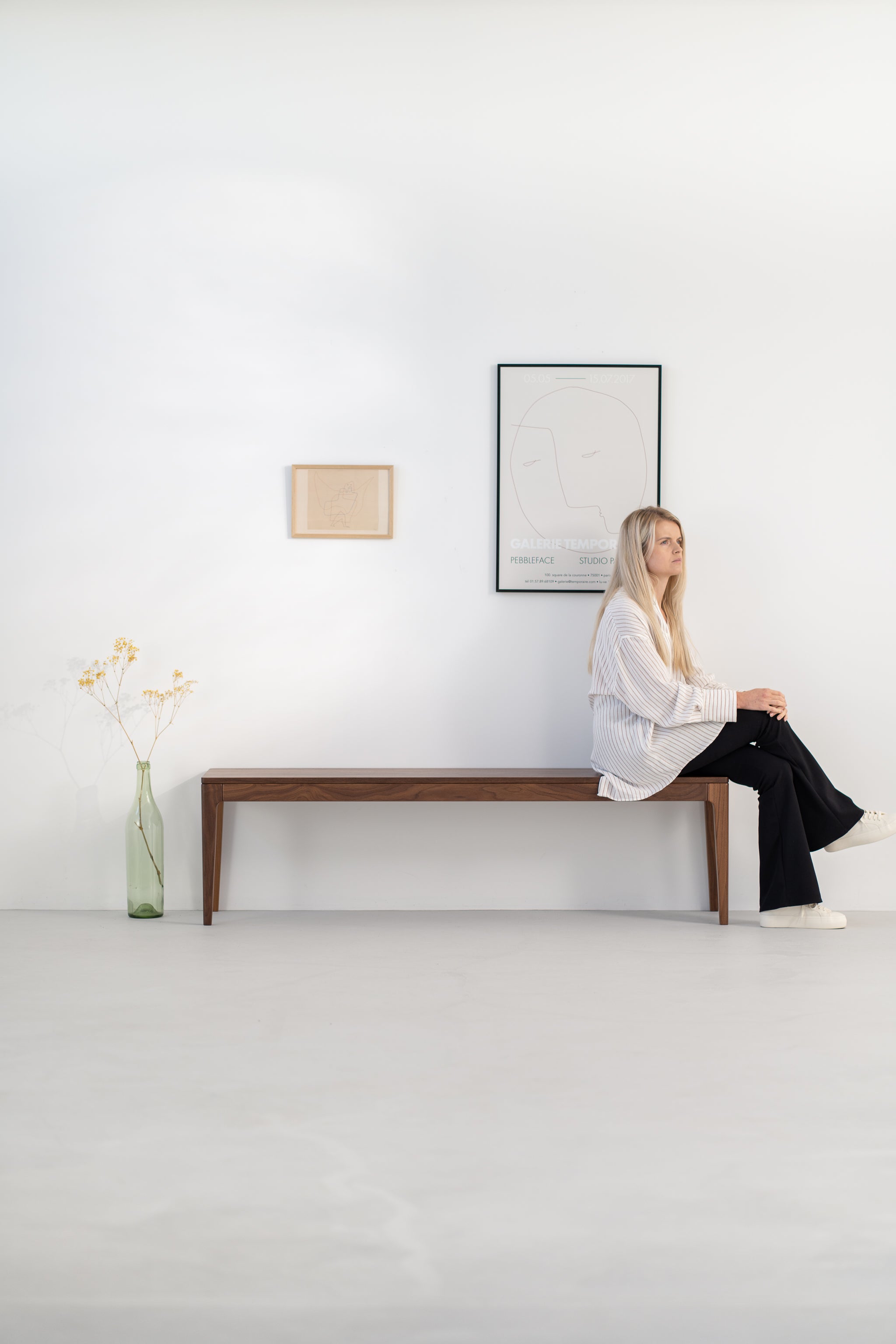 Woman sitting on a wooden bench against a minimalist white wall with framed art and a vase.