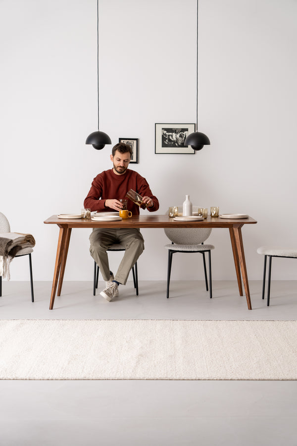 Man at a dining table pouring coffee from a French press into a mug, with modern decor and pendant lights above.