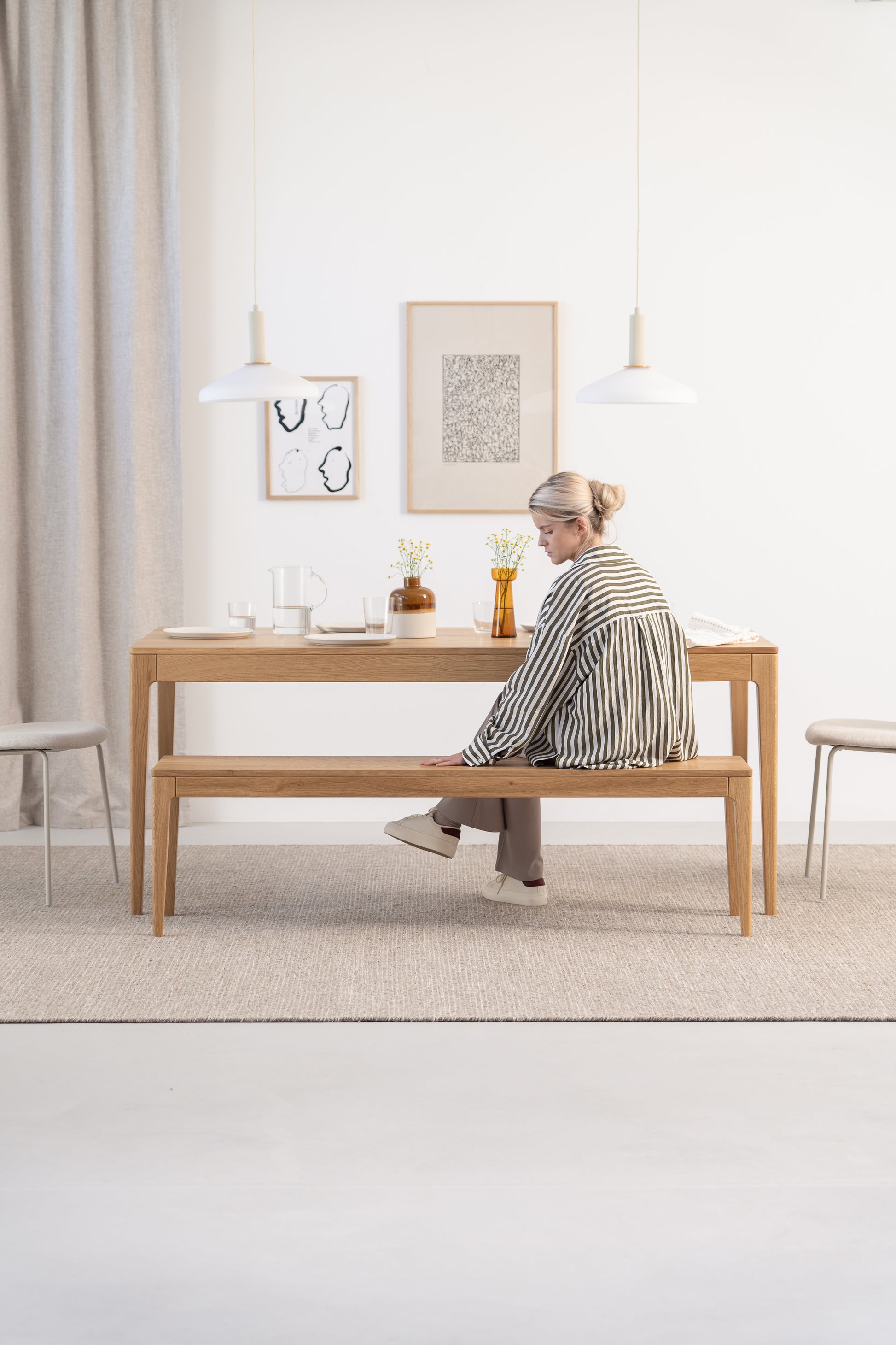 A woman sitting on a wooden bench at a minimalist dining table, with framed art and pendant lights above.