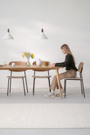 Woman in a green sweater seated at the YRKE solid oak dining table, vase of yellow flowers, two white pendant lamps above
