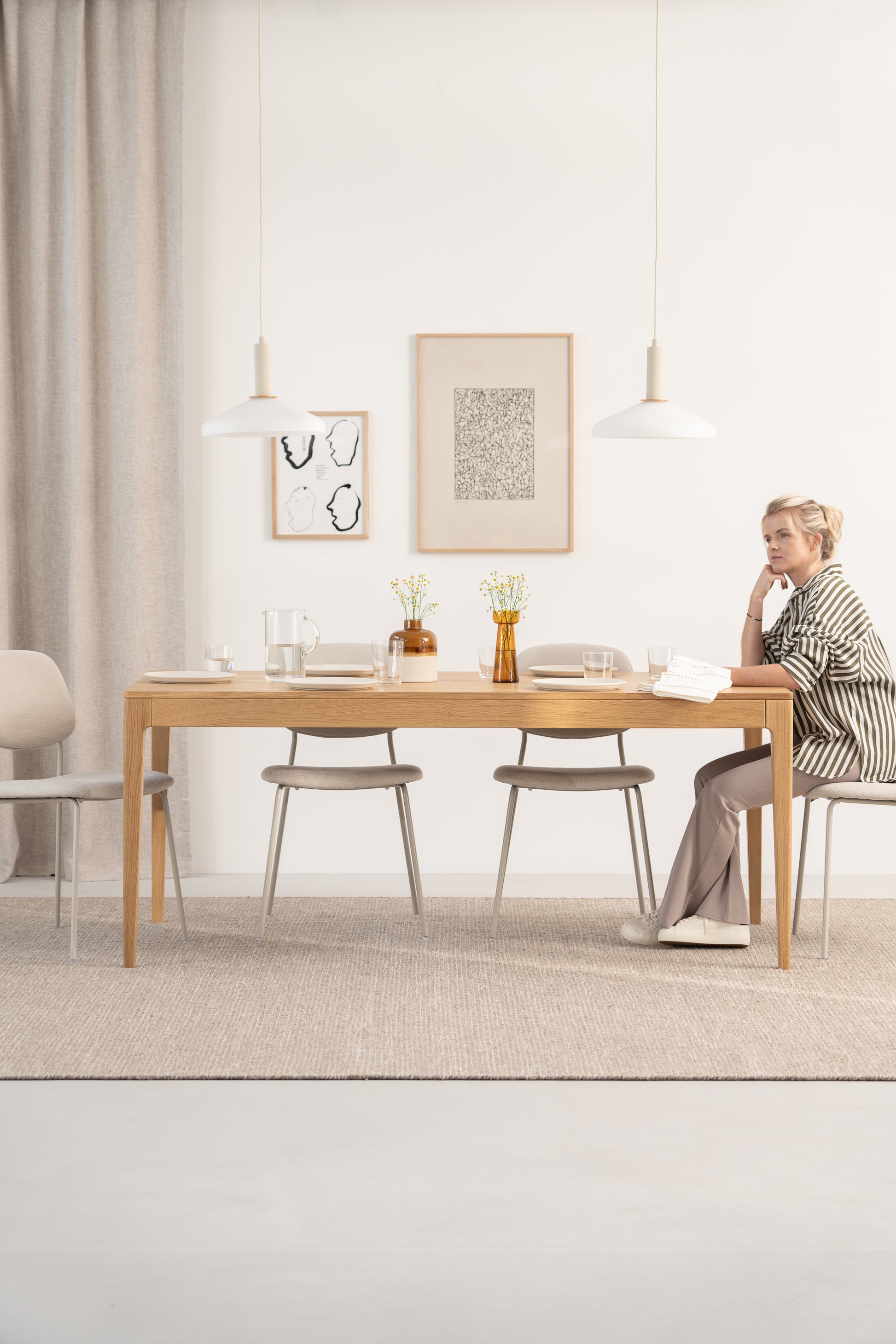 A person sits at a minimalist wooden dining table with chairs and decorations in a modern, neutral-toned room.