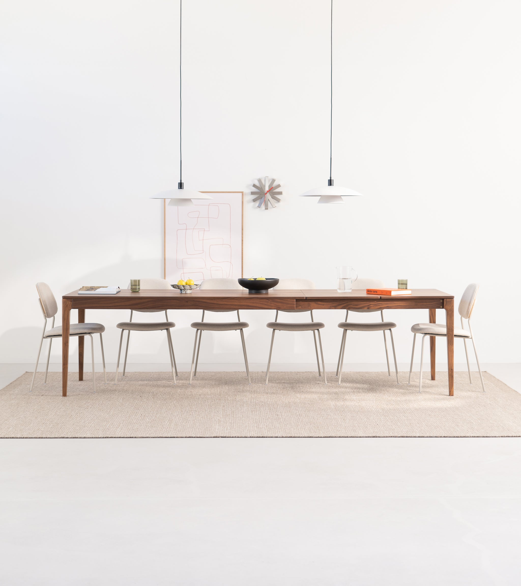Minimalist dining room with wooden table, six chairs, pendant lights, and artwork on a beige rug.