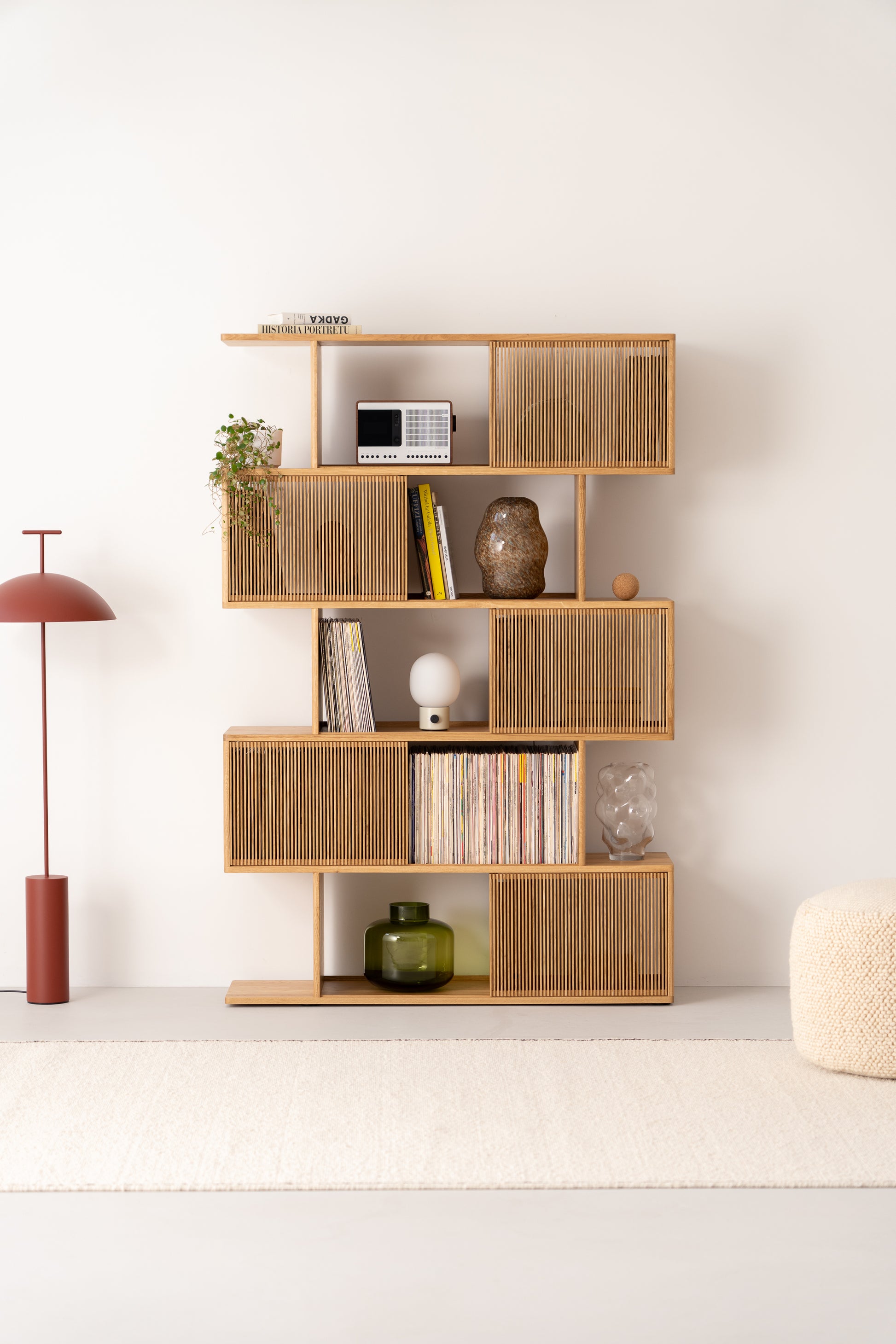 Modern wooden shelf with decor items, books, and plants. Nearby, a red floor lamp and a light ottoman on a beige rug.