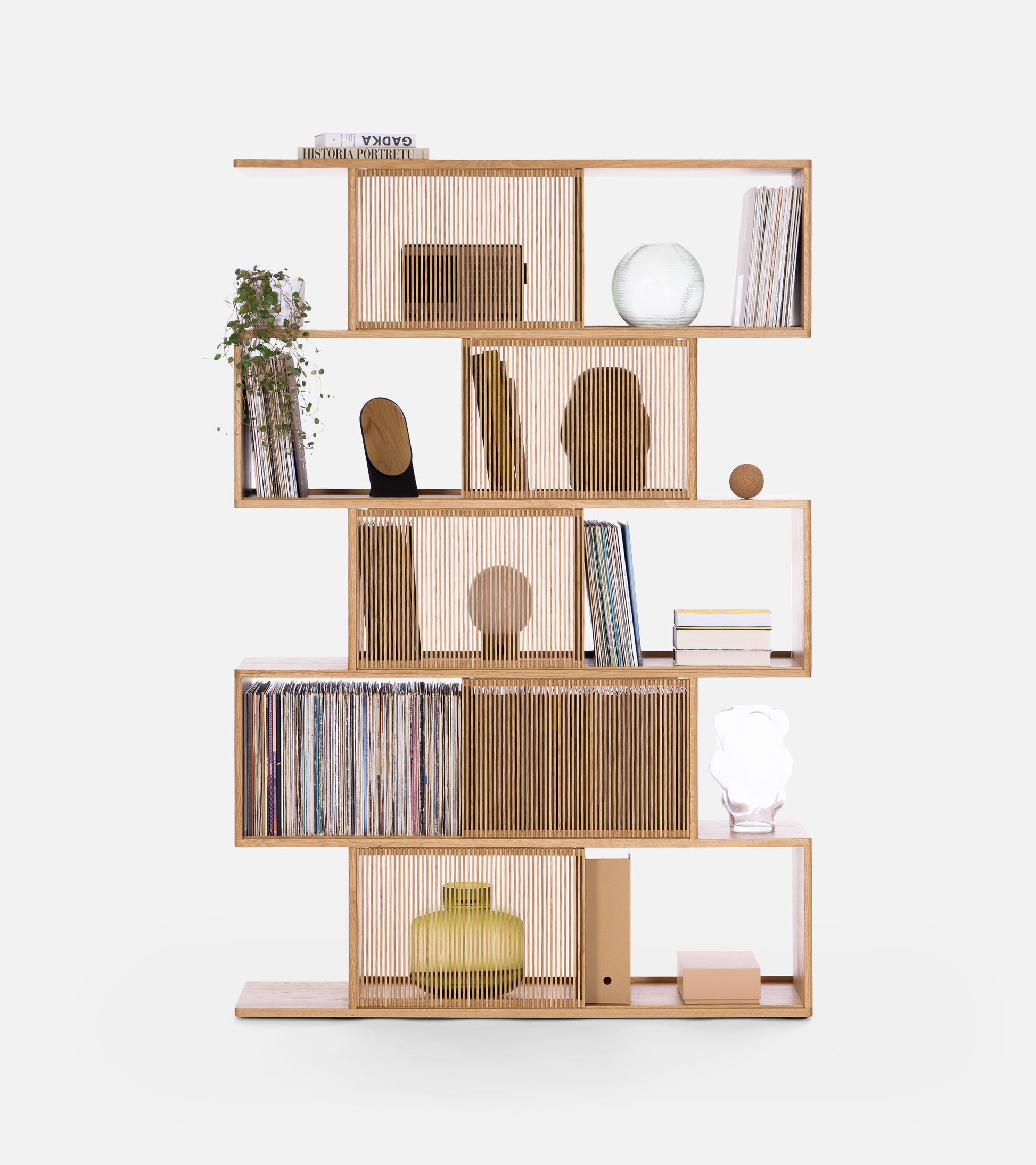 Modern wooden bookshelf with staggered shelves, books, plants, vases, and decorative items against a white background.