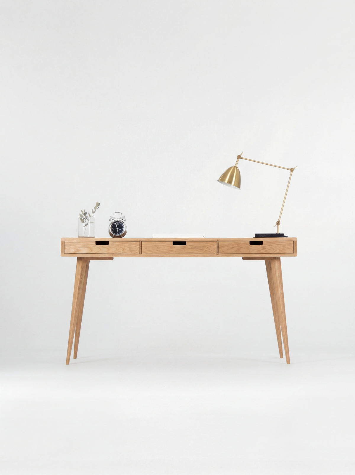 Front view of the HEIM 140 solid oak computer desk with three closed drawers, a brass lamp and alarm clock