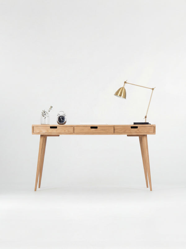 Front view of the HEIM 140 solid oak computer desk with three closed drawers, a brass lamp and alarm clock