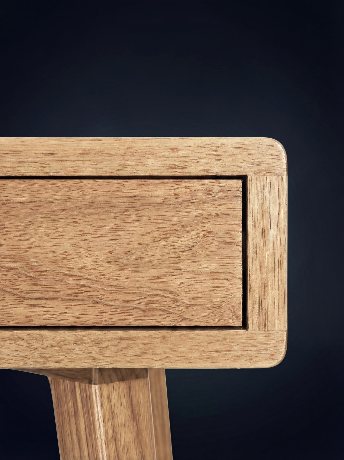 Close-up of the HEIM 105 desk in solid oak, showing a rounded body corner, drawer front and the top of a leg