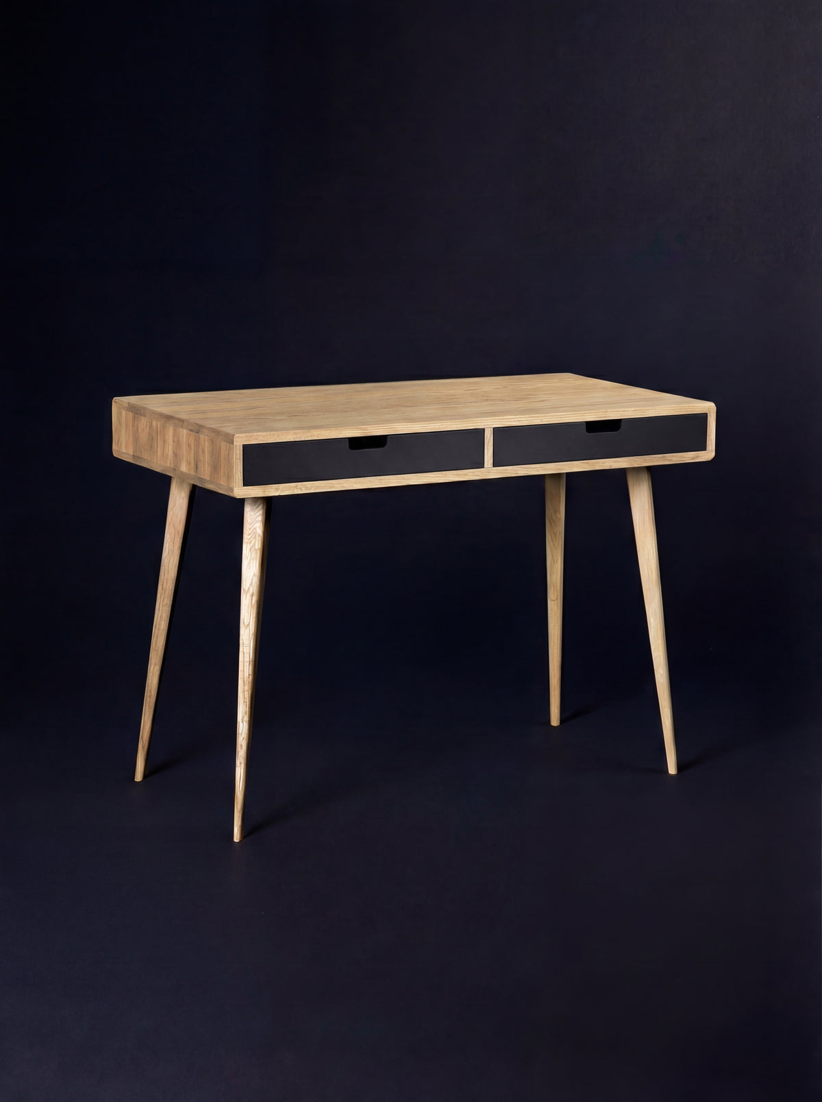HEIM 105 desk in solid oak, seen at an angle with a bare top and two black drawer fronts on a dark background