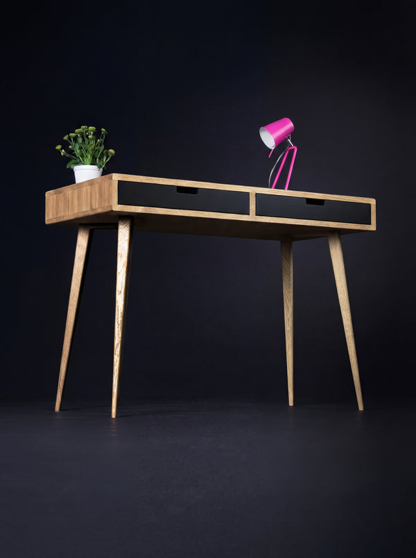 Angled view of the HEIM 105 desk in solid oak with two black drawer fronts, a pink lamp and a plant