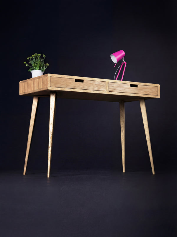 HEIM 105 desk in solid oak at a three-quarter angle on a dark background, with a pink lamp and plant on top