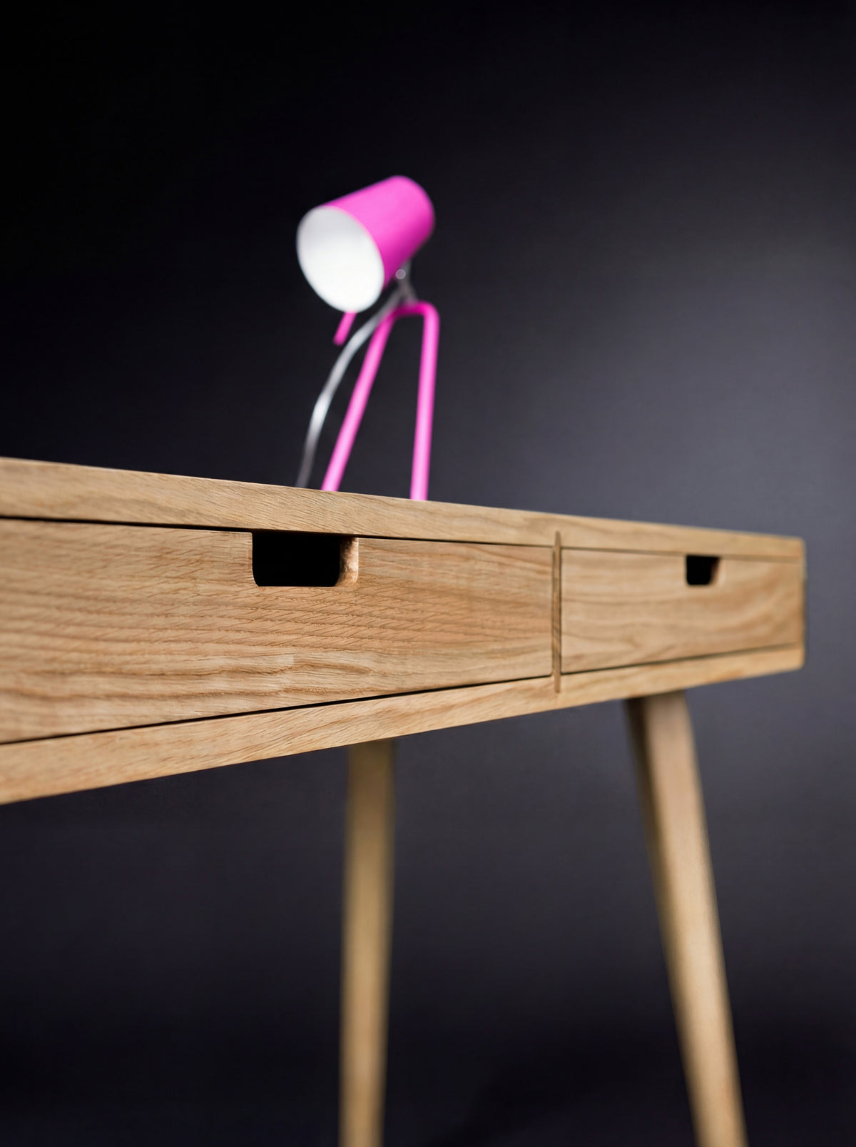 HEIM 105 in solid oak, close-up along two drawer fronts with rectangular cutout grips and a blurred pink lamp above