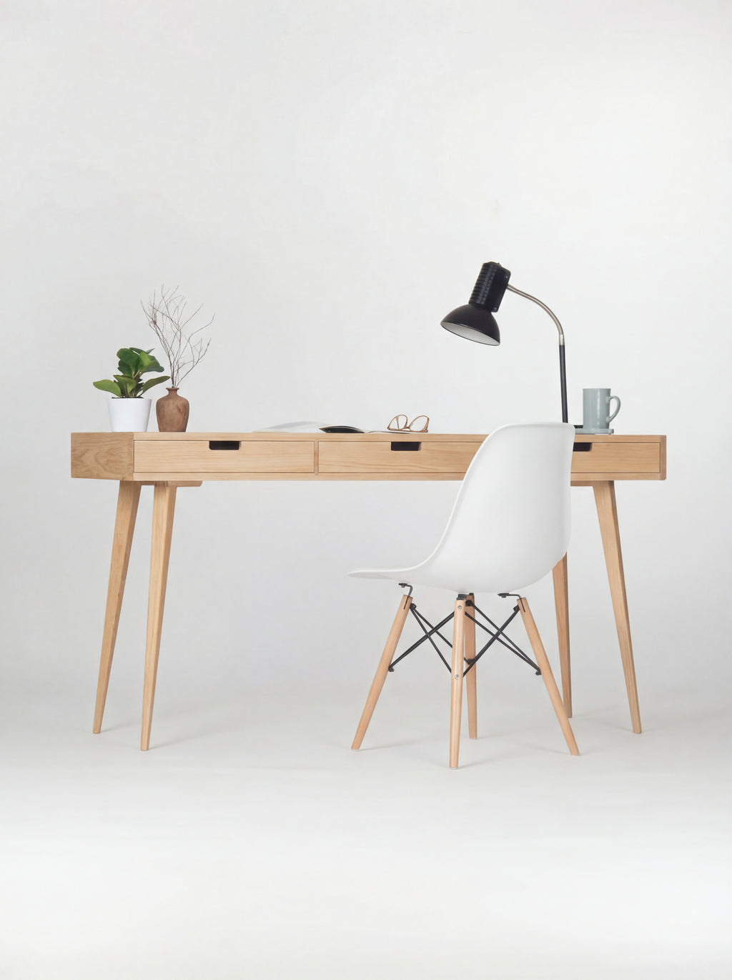 Front view of the HEIM 140 oak wood desk with a white shell chair, black lamp, plant and vase with twigs