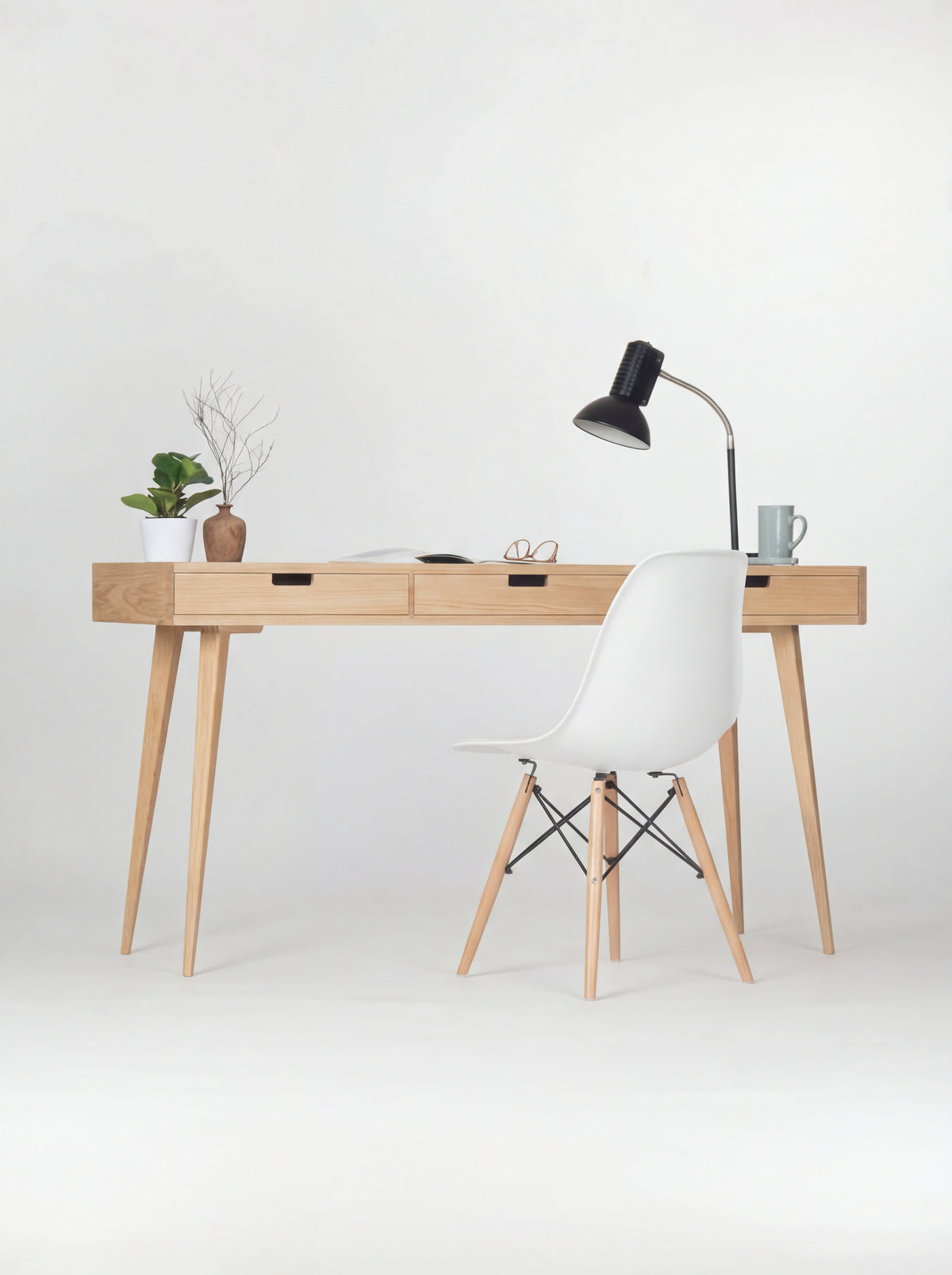Front view of the HEIM 140 oak wood desk with a white shell chair, black lamp, plant and vase with twigs