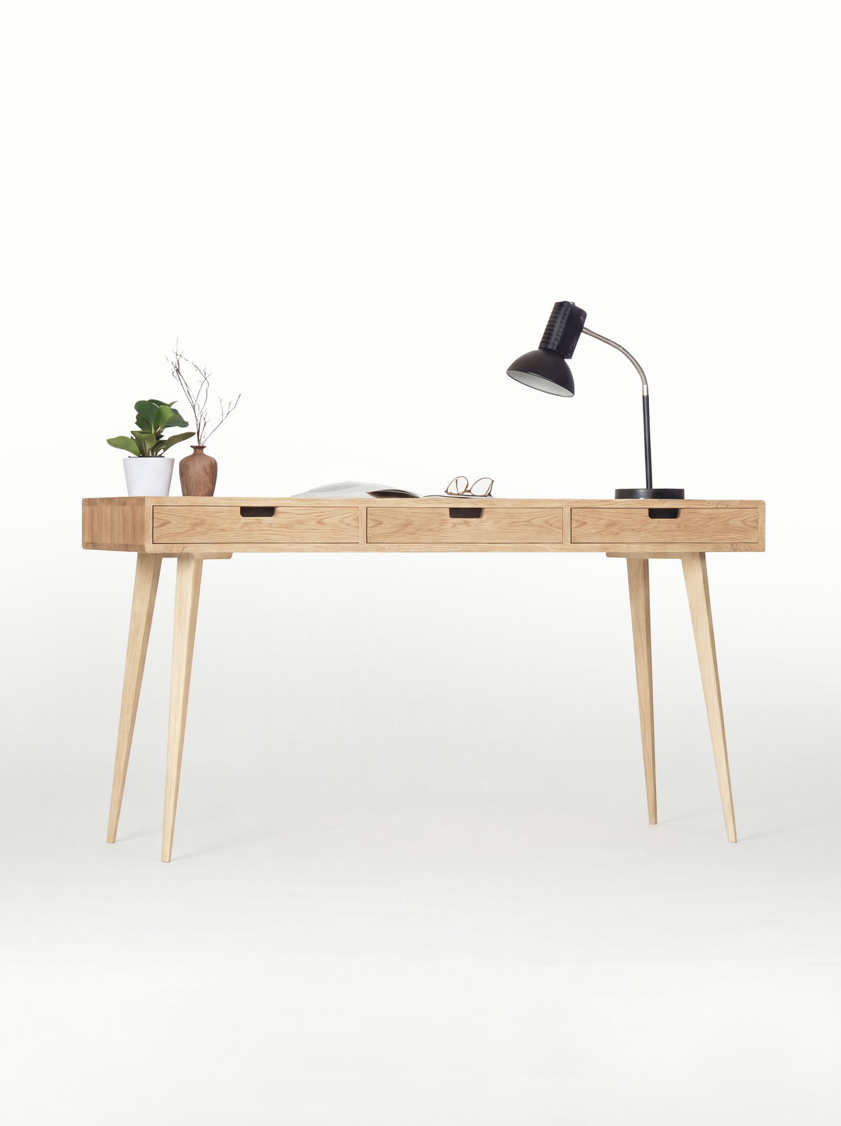 HEIM 140 office desk in solid oak, front view with natural oak drawer fronts, a black lamp and an open magazine