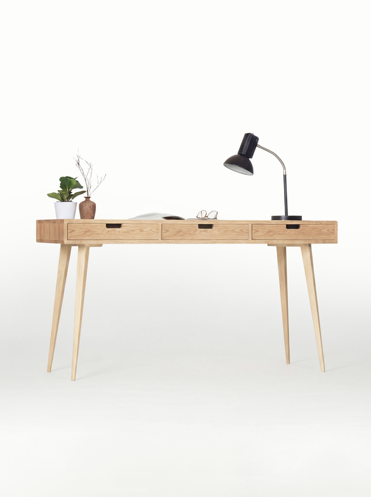 HEIM 140 office desk in solid oak, front view with natural oak drawer fronts, a black lamp and an open magazine