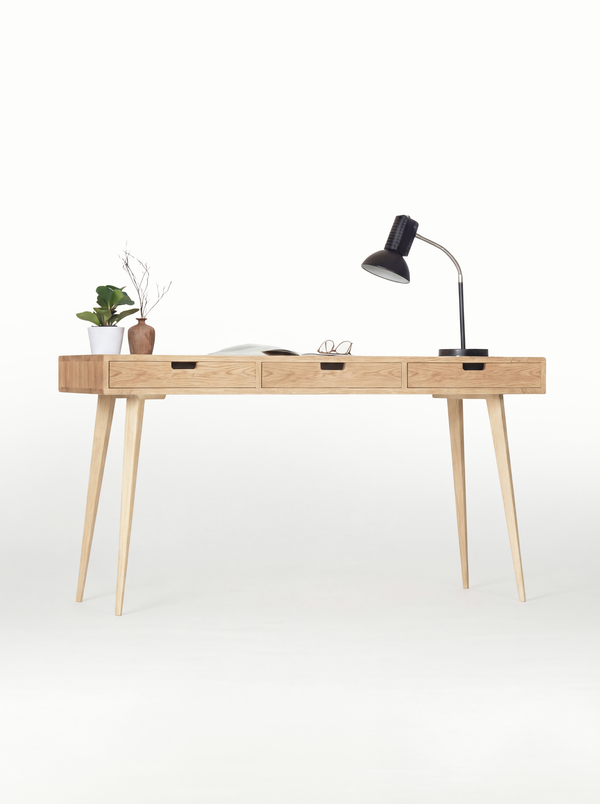 HEIM 140 office desk in solid oak, front view with natural oak drawer fronts, a black lamp and an open magazine