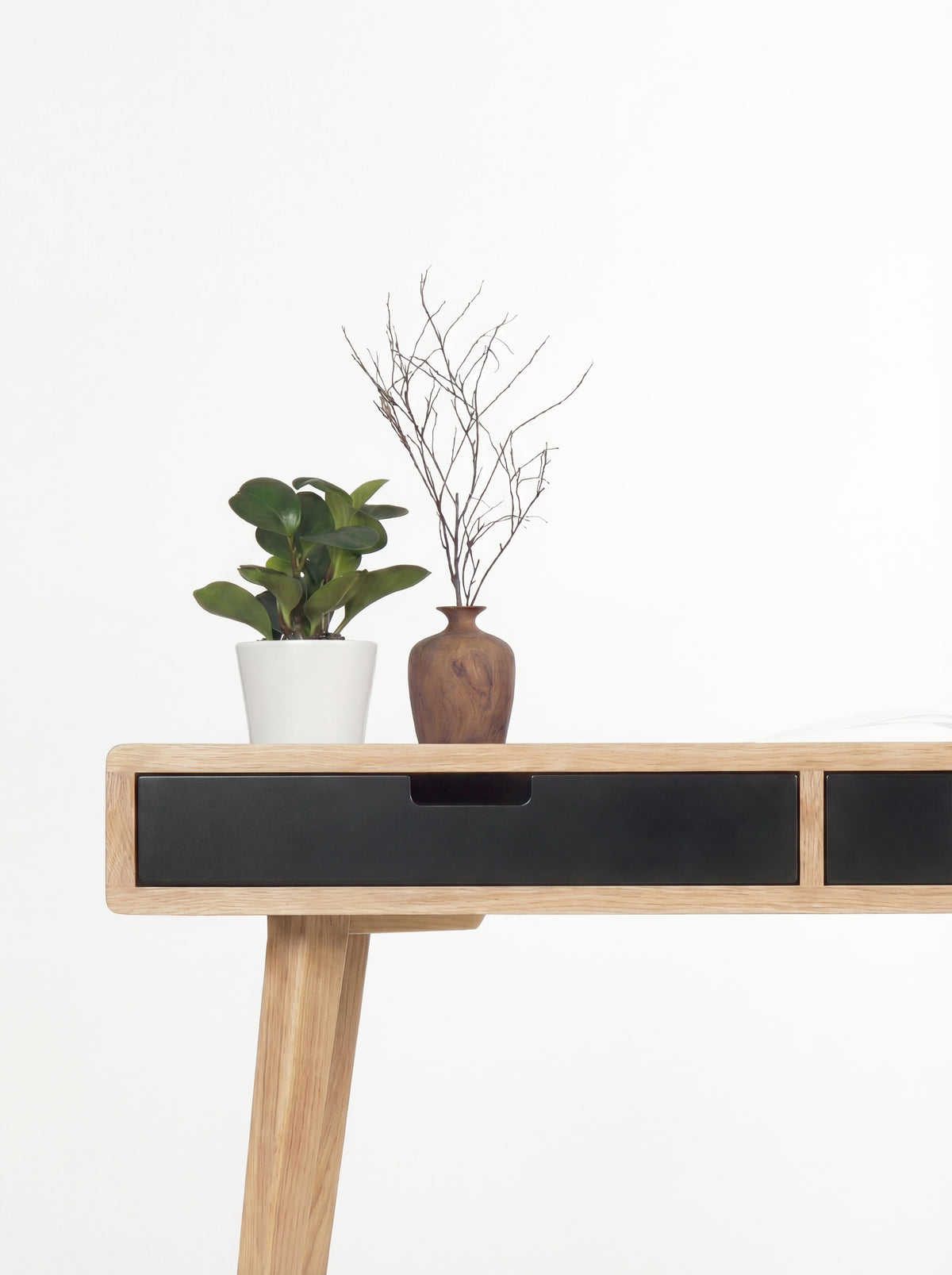 Close-up of the HEIM 140 desk in solid oak, with a black drawer front, cut-out grip and wooden vase with twigs