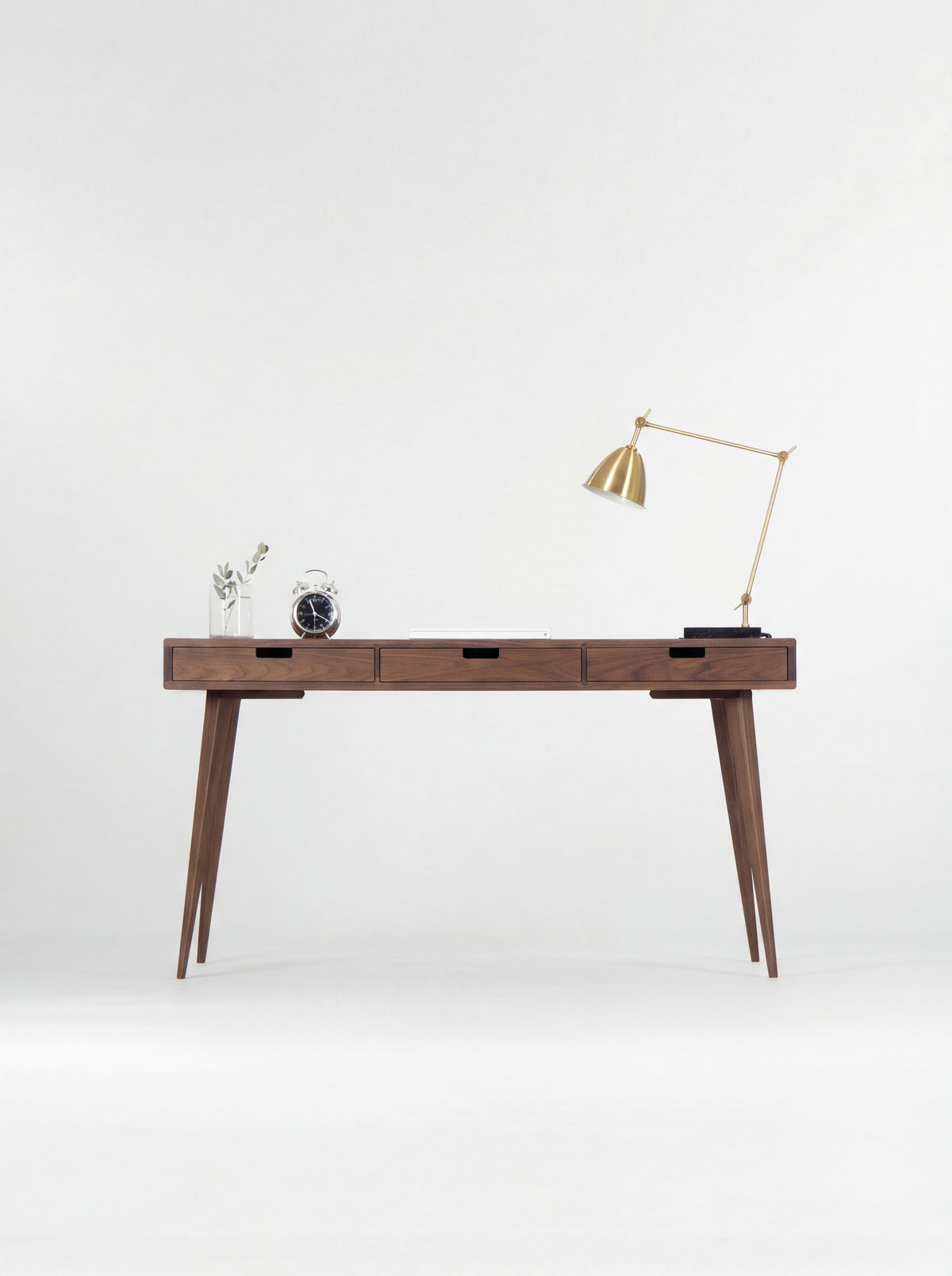 HEIM 140 desk in solid walnut, straight-on view of three closed drawers with cut-out grips, a brass lamp and clock