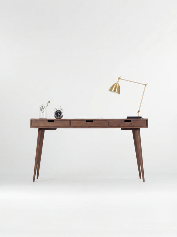 HEIM 140 desk in solid walnut, straight-on view of three closed drawers with cut-out grips, a brass lamp and clock