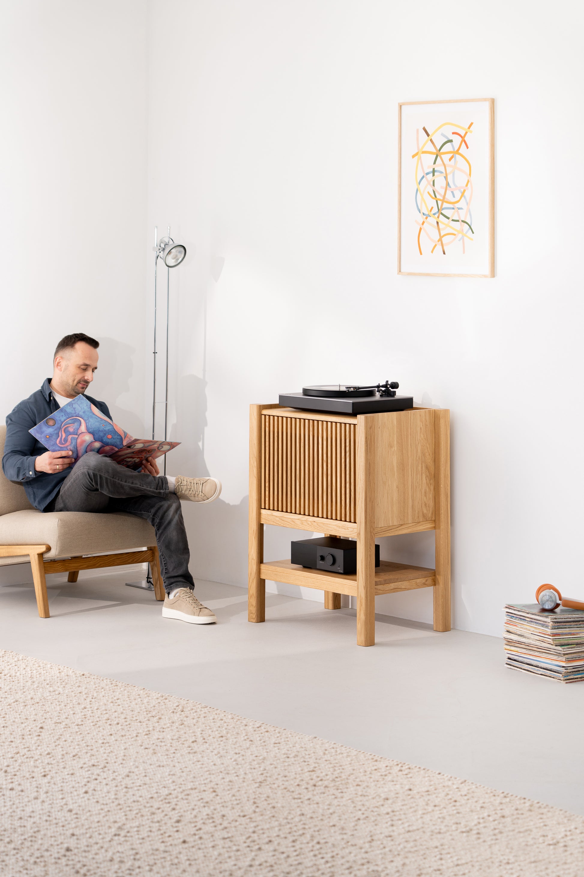 Man sitting, reading a vinyl cover beside a wooden cabinet with a record player, in a minimalist room.