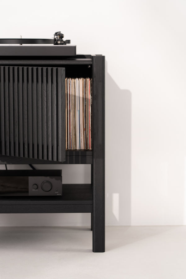 Black cabinet with a turntable, vinyl records, and audio equipment against a white wall.