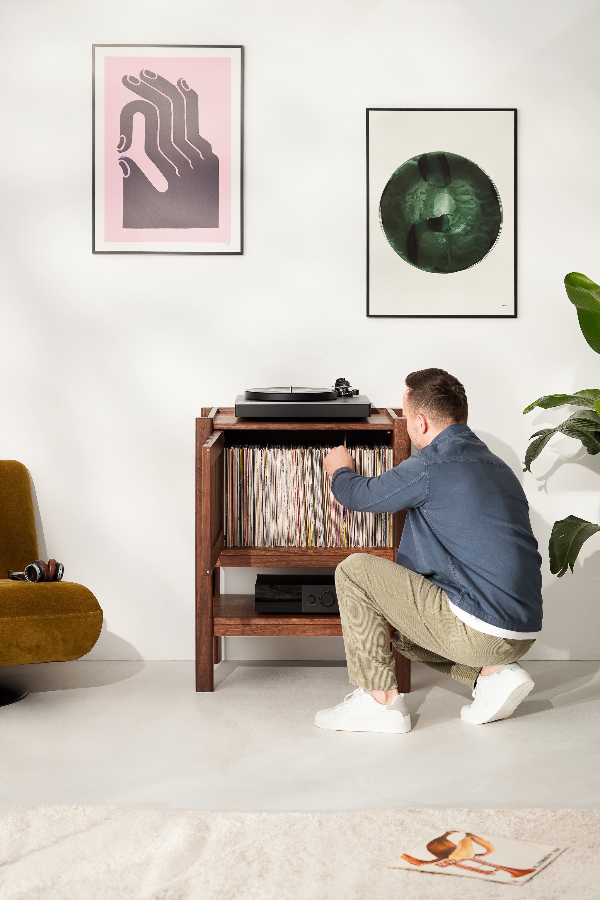 Man selecting a vinyl record from a wooden cabinet, with artwork on the wall and a turntable on top.