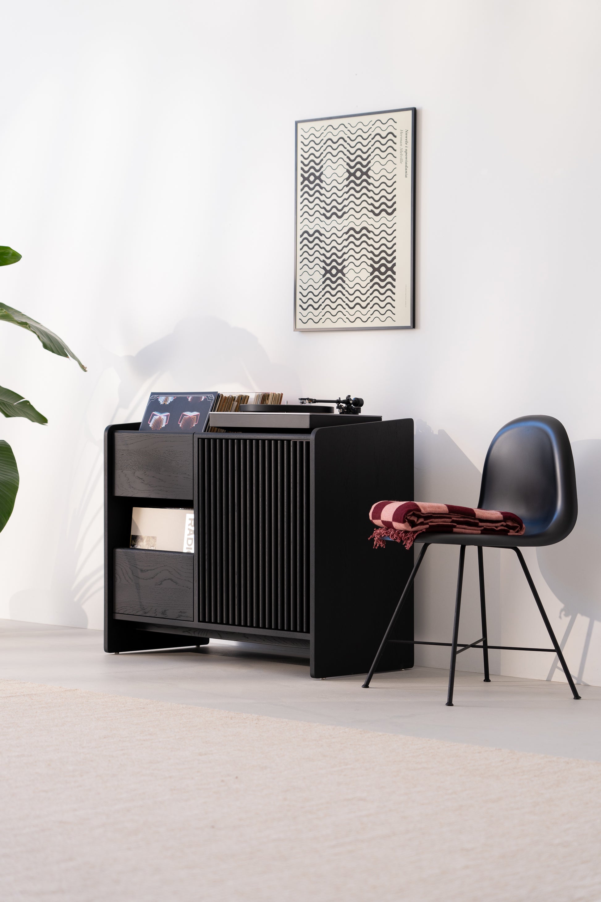 Black cabinet with records, abstract art, chair with blanket, and a plant. Minimalist, modern interior design.