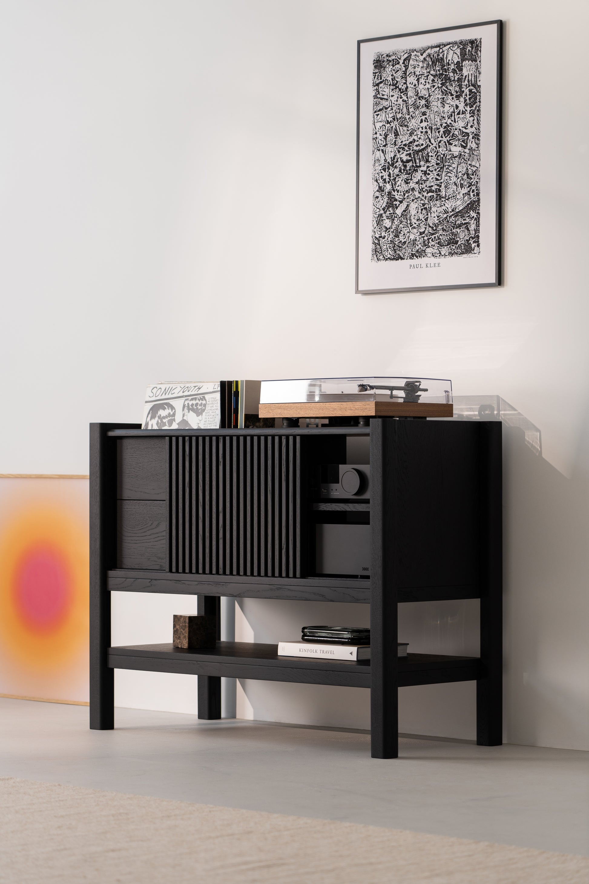 Black cabinet with a turntable and records, under a framed artwork. A colorful print leans against the wall.