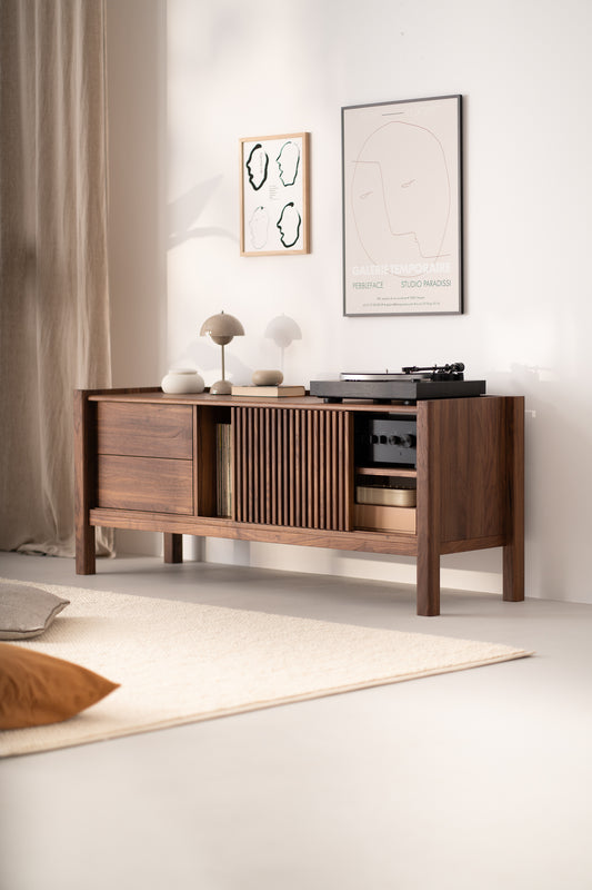 A modern wooden sideboard with lamps, books, and a record player, with abstract wall art above, in a minimal living room.