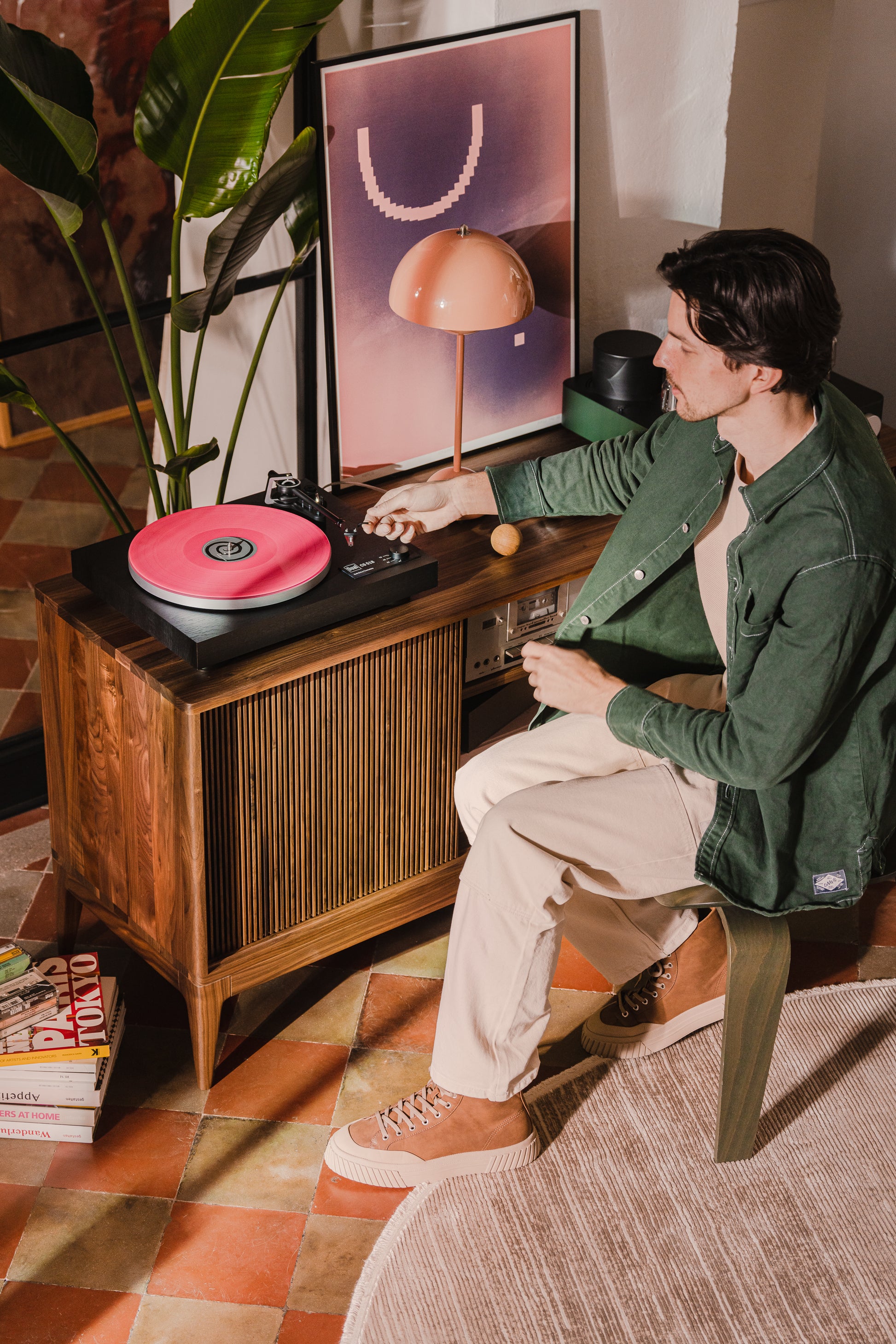 A person in a green jacket plays a pink vinyl record on a wooden turntable in a cozy room.