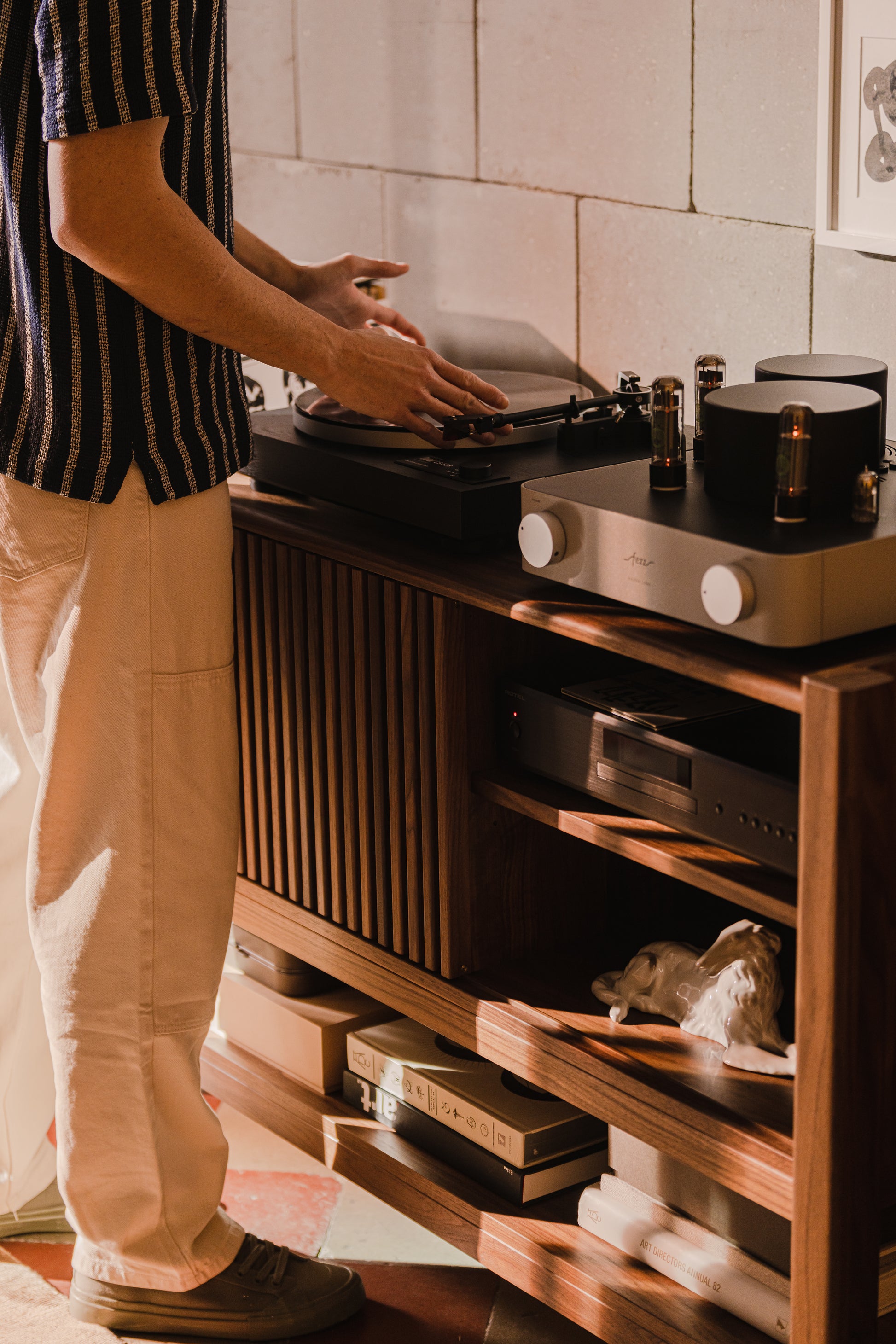 Person adjusting a turntable next to audio equipment on a wooden cabinet.