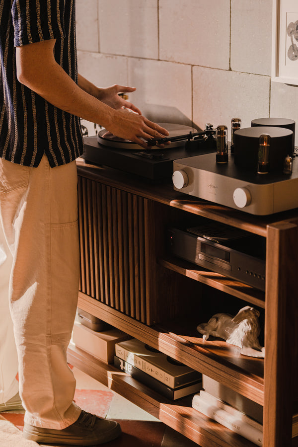 Person adjusting a turntable next to audio equipment on a wooden cabinet.