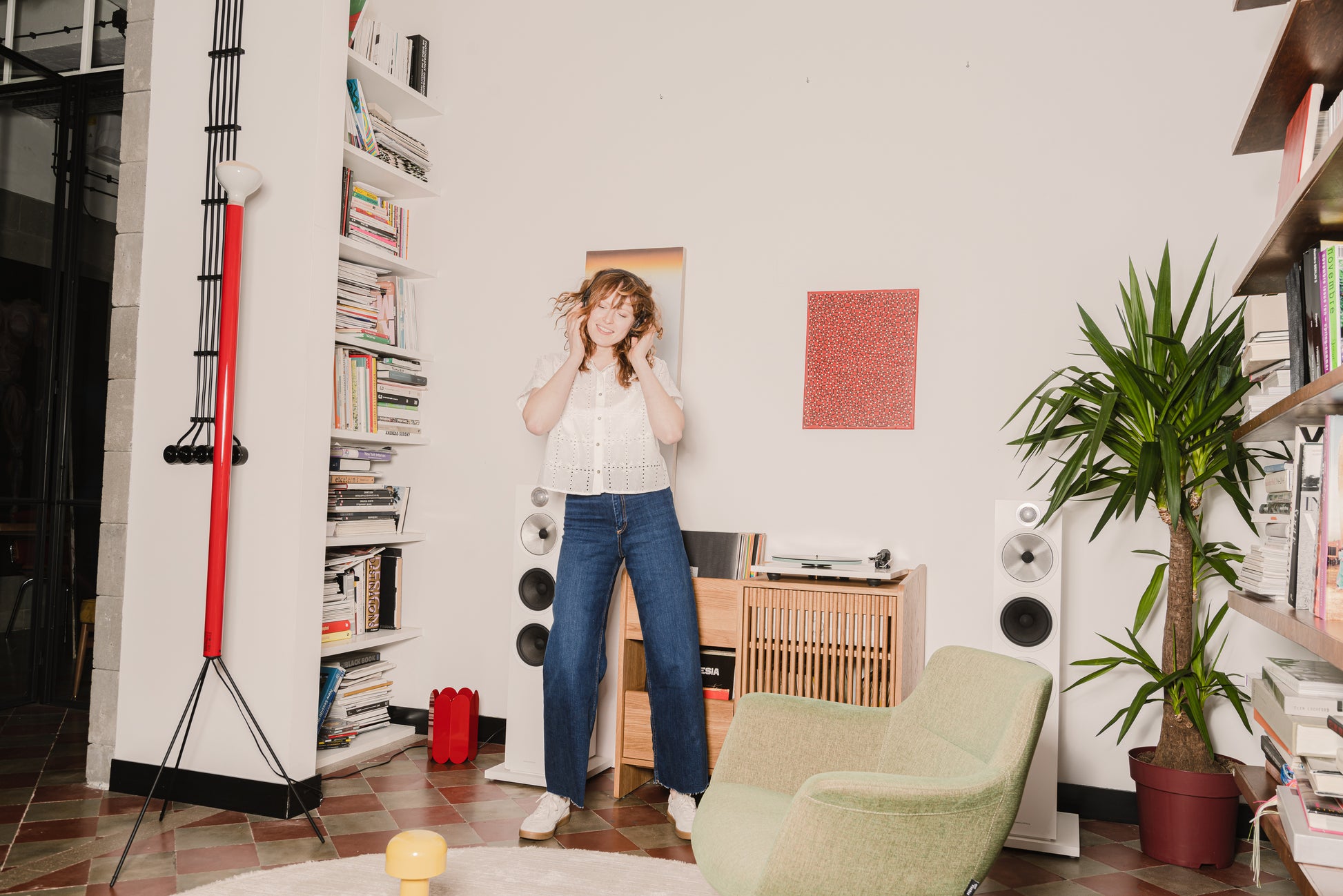 Woman with curly hair enjoys music in a cozy, book-filled living room with speakers and a plant.