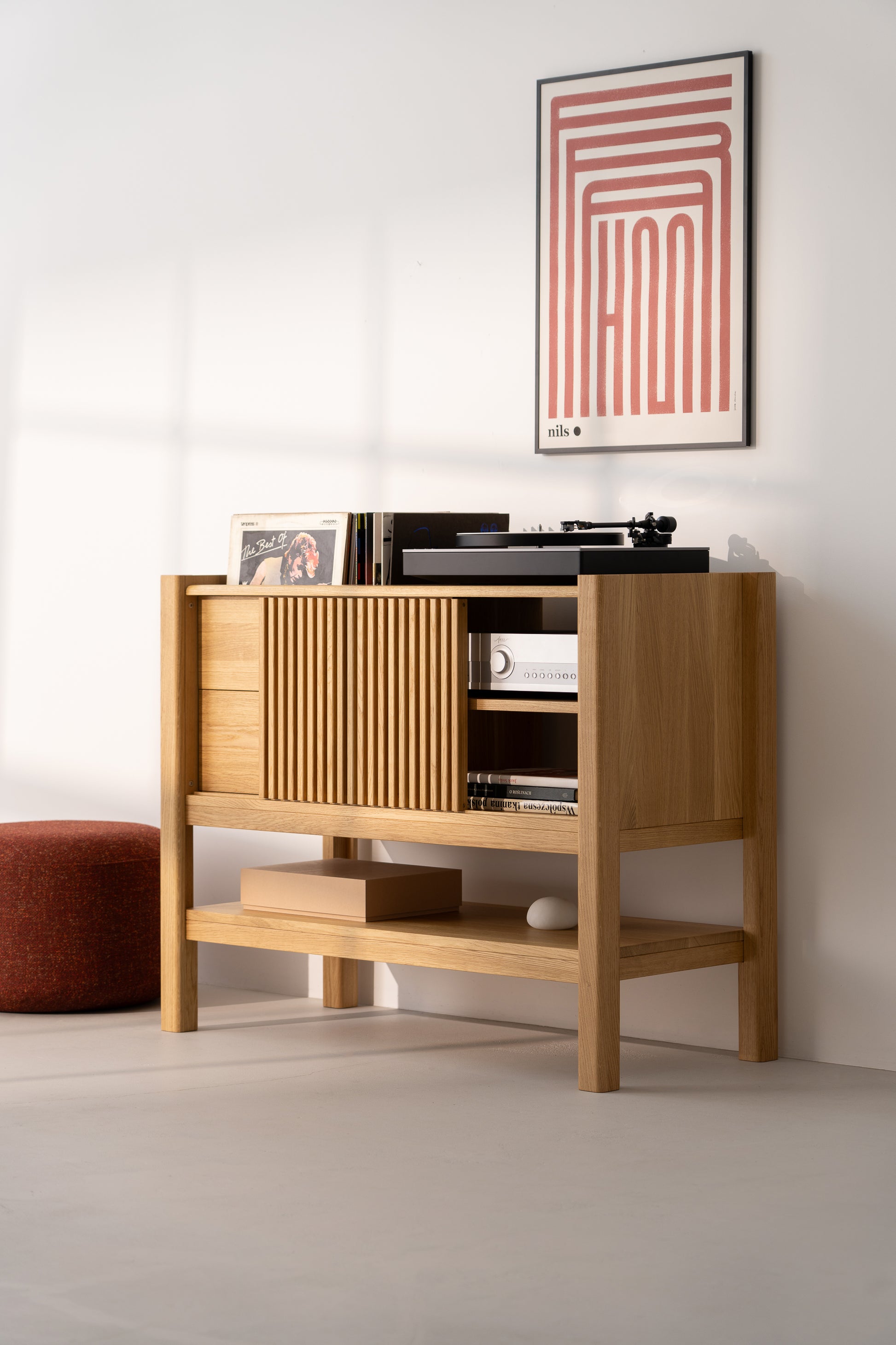 A cabinet for a turntable and vinyl records stands against a white wall, with a poster hanging above it and a pouf next to it.