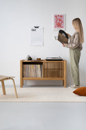 Woman holding a vinyl record beside the JAMM LOW 111 solid oak stand, records in the open compartment, turntable on top
